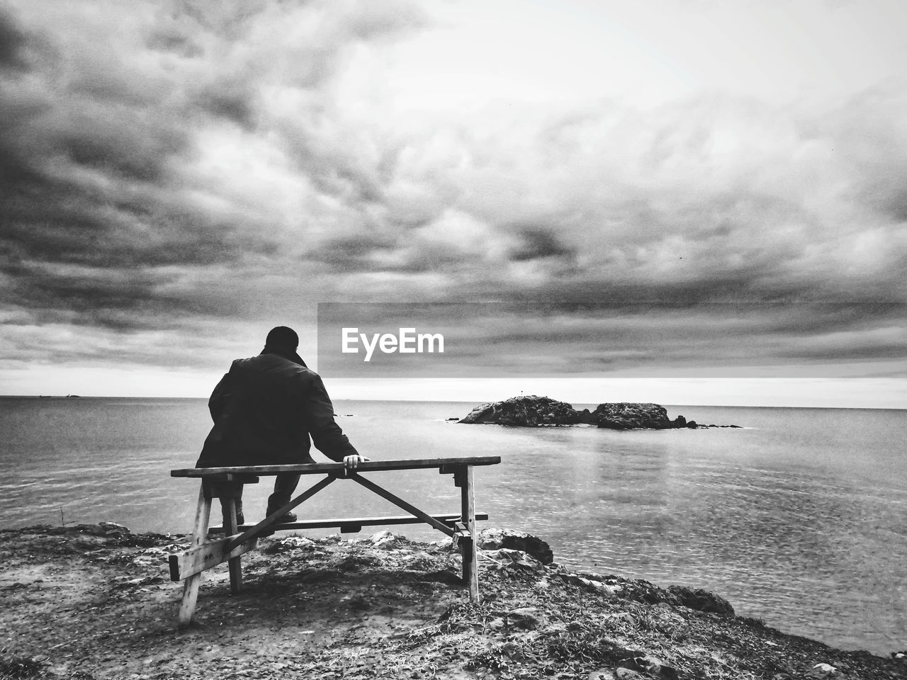 REAR VIEW OF MAN SITTING ON SHORE AGAINST SKY