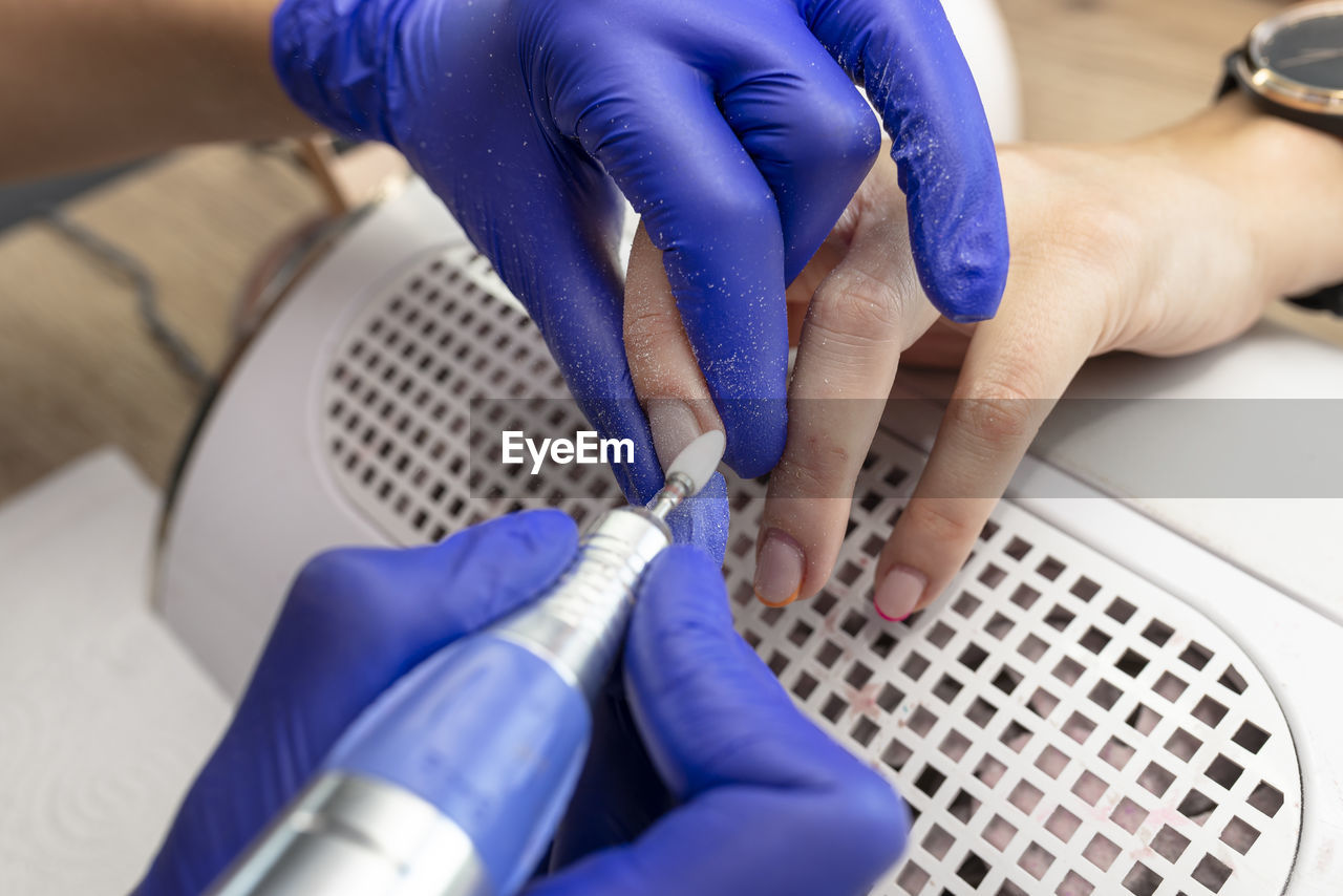 The manicurist grinds the nail plate with a milling machine in blue latex gloves.