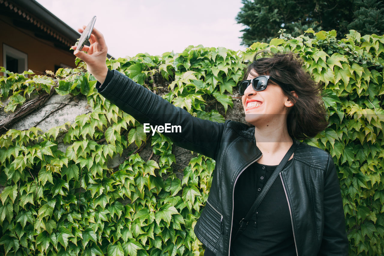 Smiling woman taking selfie with smart phone while standing against plants at park