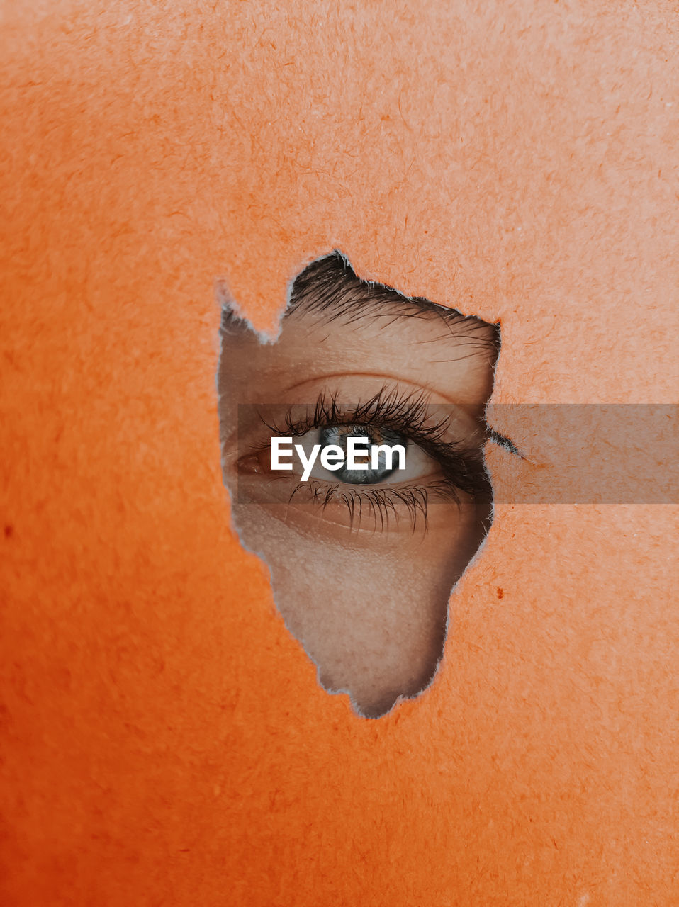 Close-up eye peeking through torn orange paper