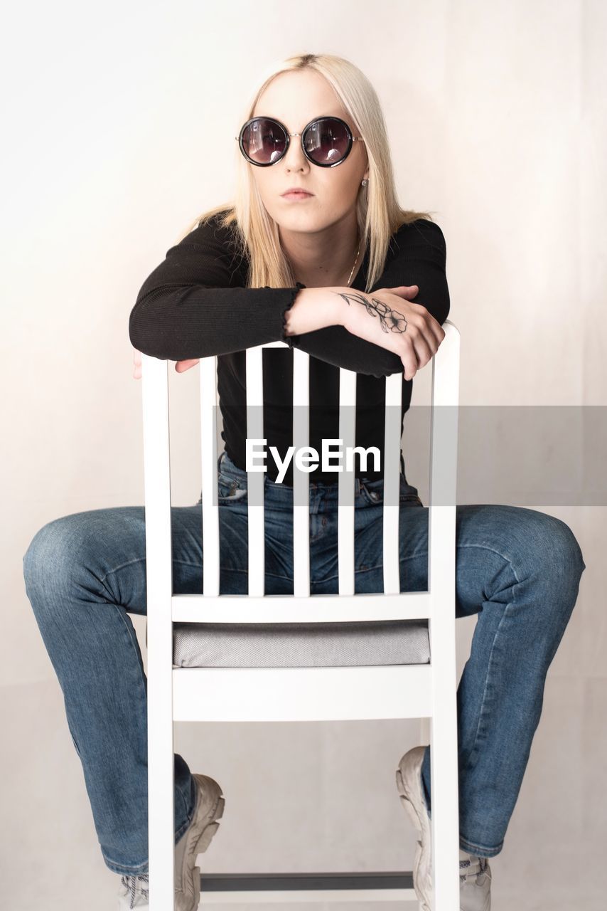 Portrait of woman wearing sunglasses while sitting on chair against wall