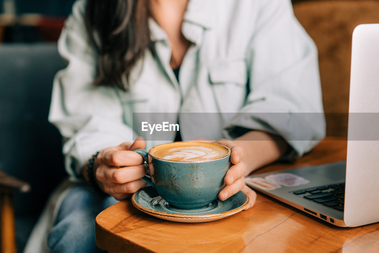 Unrecognizable cropped woman drinking coffee while working on a laptop in a cafe.