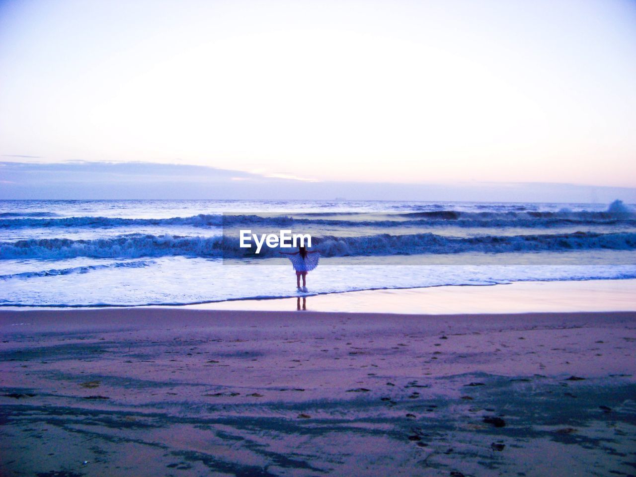 REAR VIEW OF MAN STANDING ON BEACH AGAINST SKY