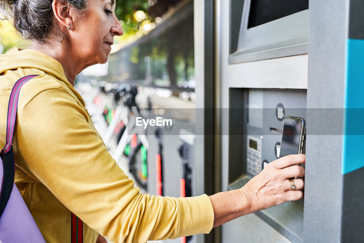 Crop senior female using smartphone to gain access to atm near electric bike rental station on city street