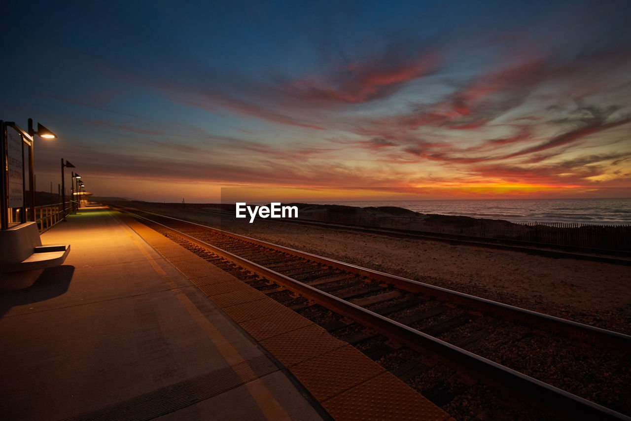 Railroad tracks by illuminated platform against sky during sunset