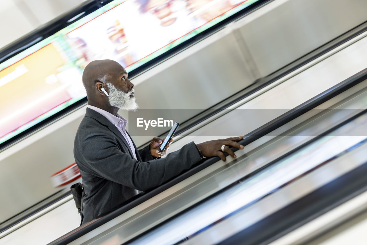 Senior businessman with smart phone moving up on escalator at subway station