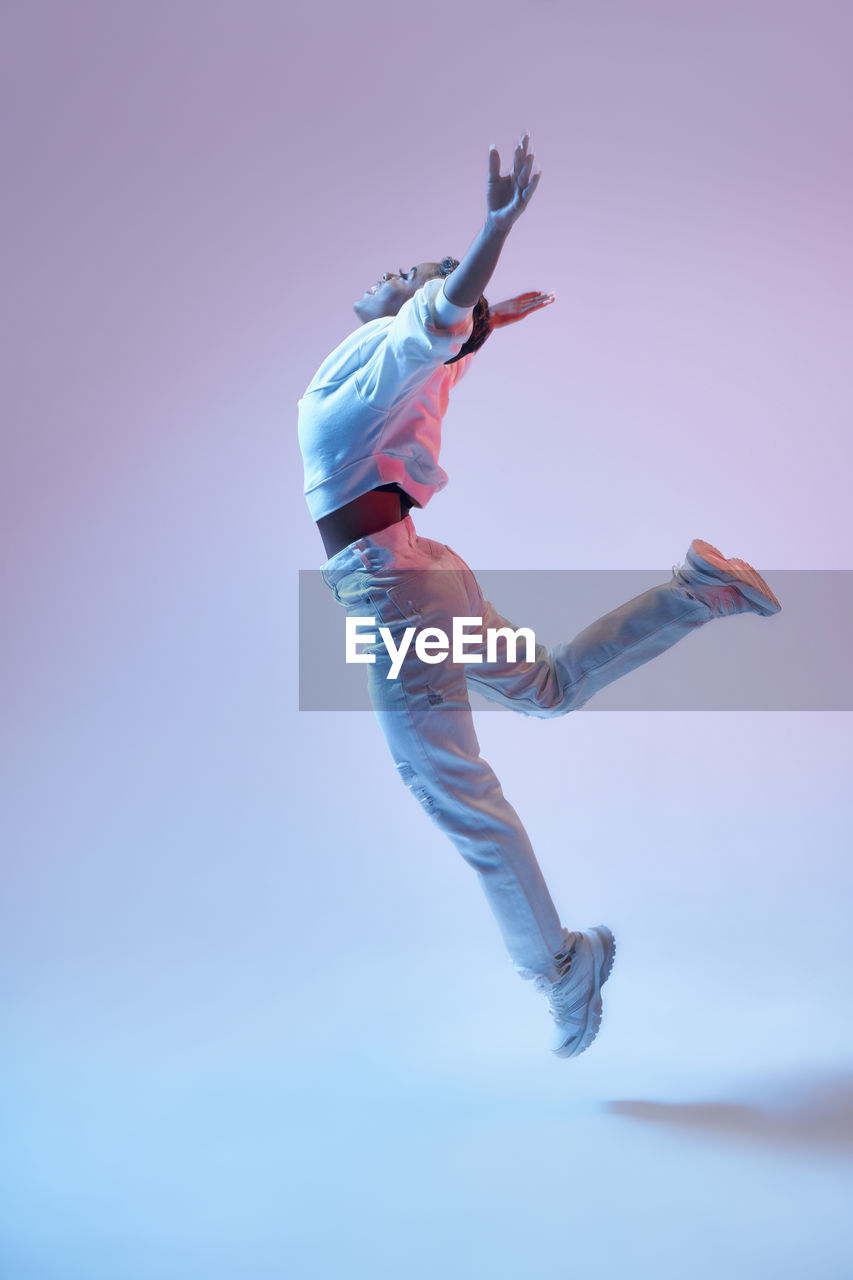 Side view of energetic ethnic woman in wireless headphones and trendy clothes jumping with open arms while dancing hip hop