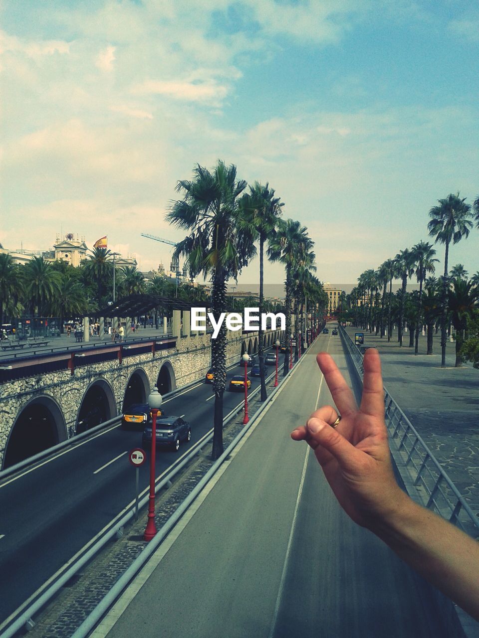 Cropped hand of person showing peace sign over road against sky