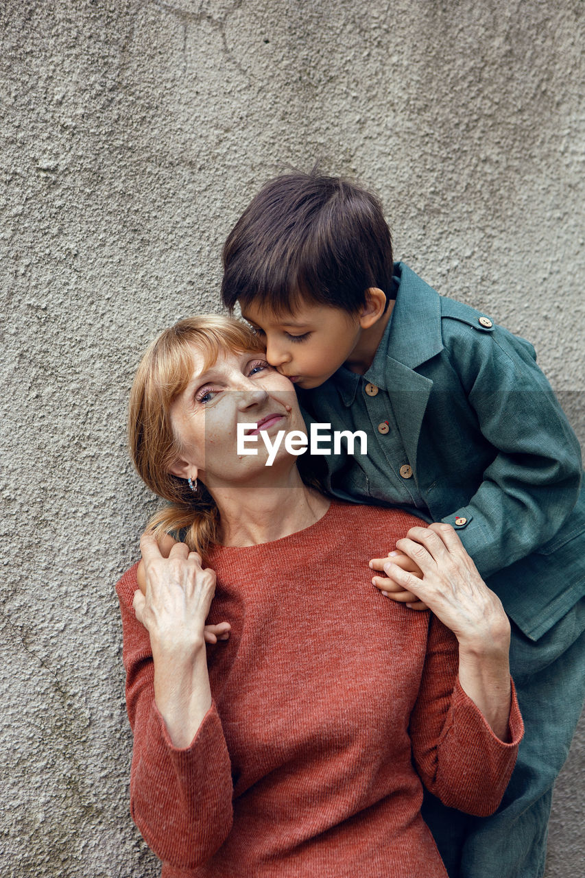 Child boy hugs his grandmother an elderly woman on the street against a gray wall