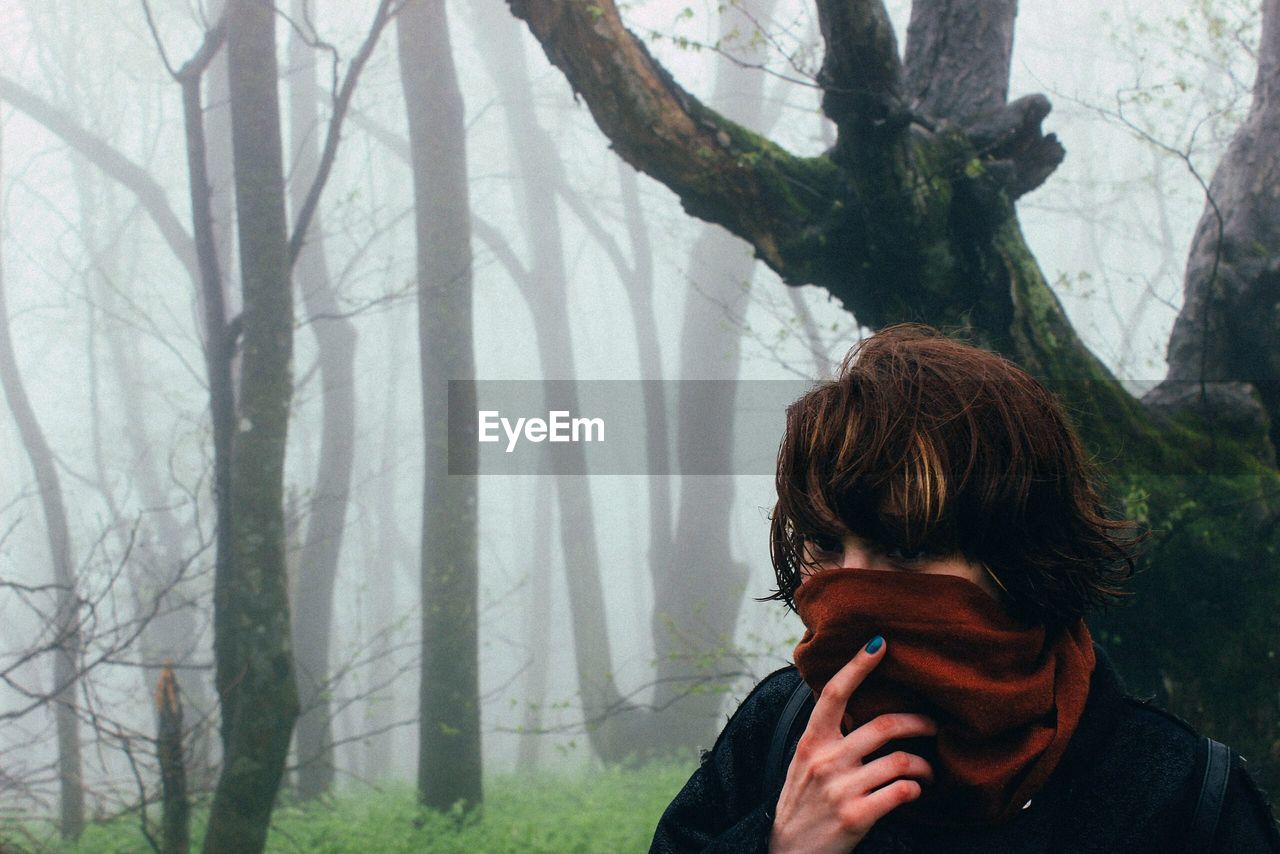 Close-up of woman with covered face against bare trees in foggy weather