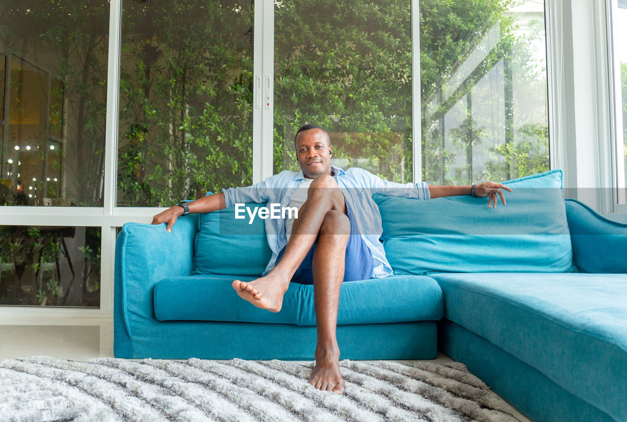 FULL LENGTH PORTRAIT OF A YOUNG MAN SITTING ON SOFA AT WINDOW