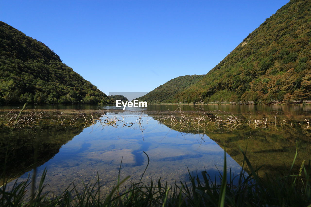 Green mountains by calm lake against clear blue sky