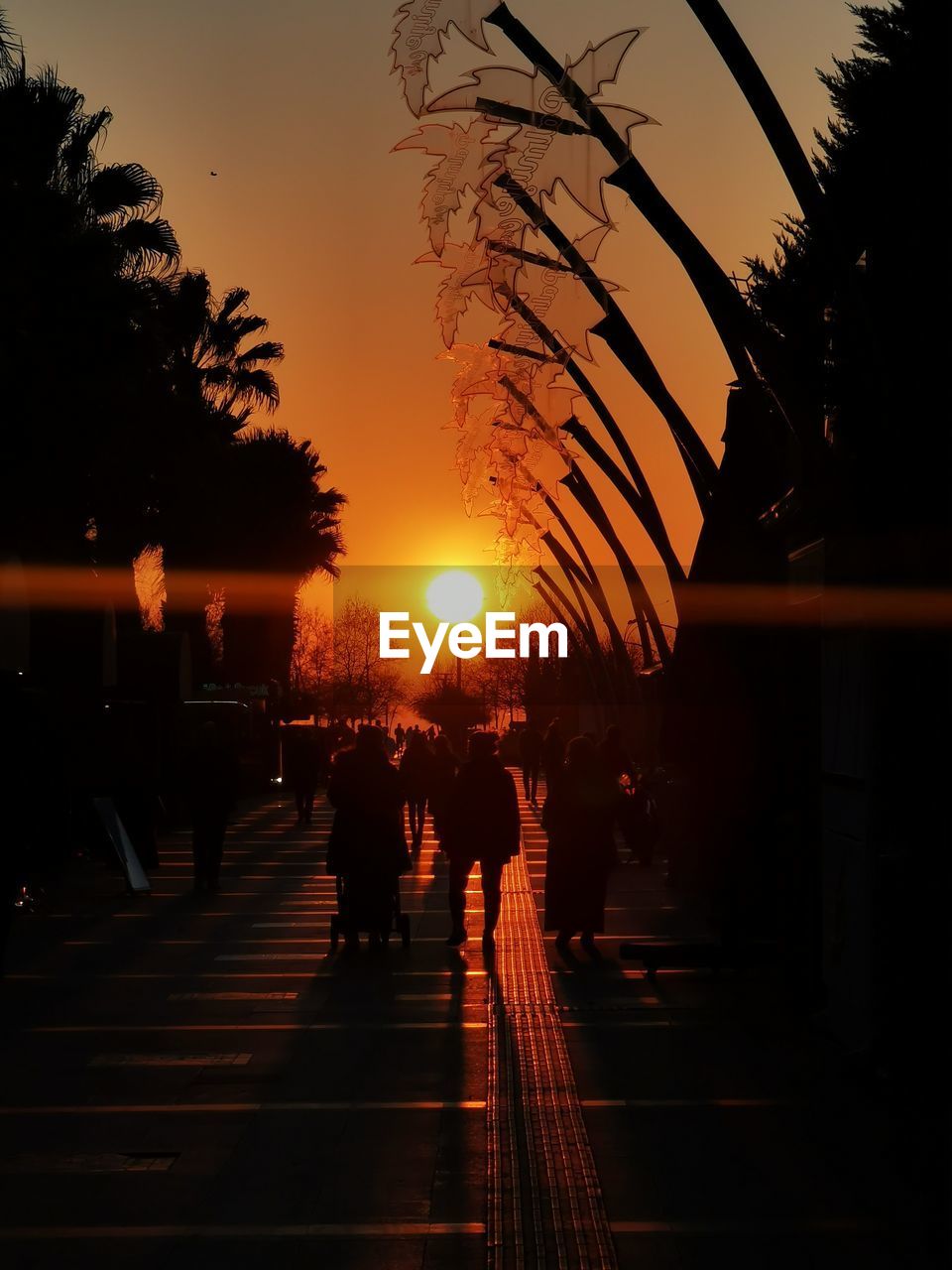 Silhouette people walking on illuminated street against orange sky