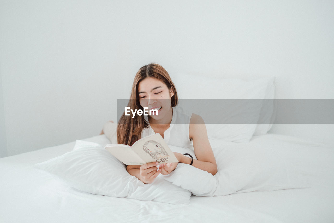 Smiling young woman reading book while lying on bed