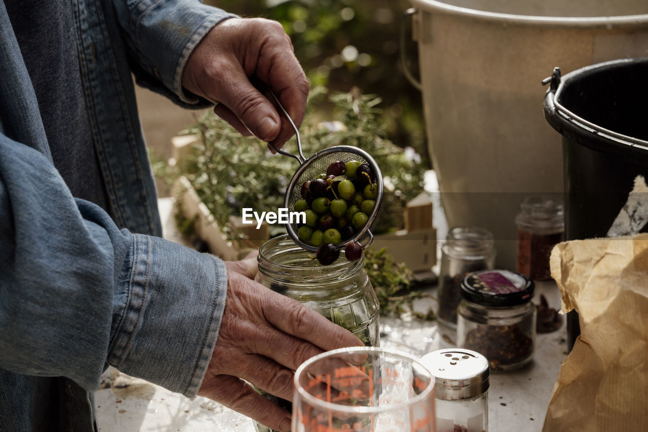 Senior man filling glass jar with olives from colander on table