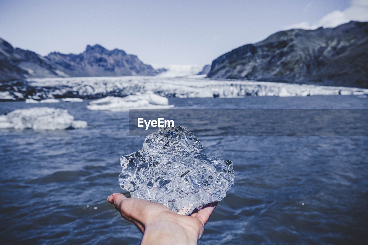 Cropped hand of person holding ice over sea against mountains and sky