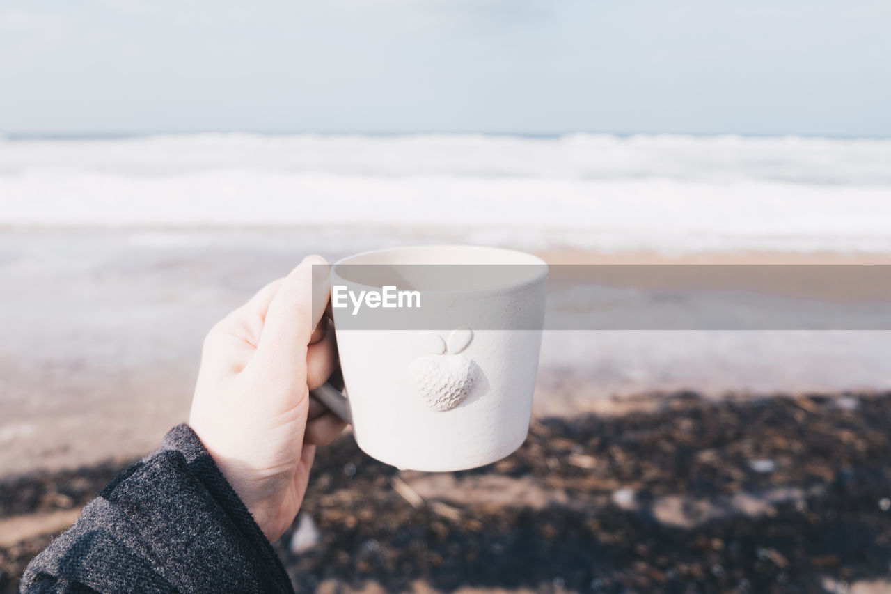 Cropped hand holding coffee cup at beach against sky