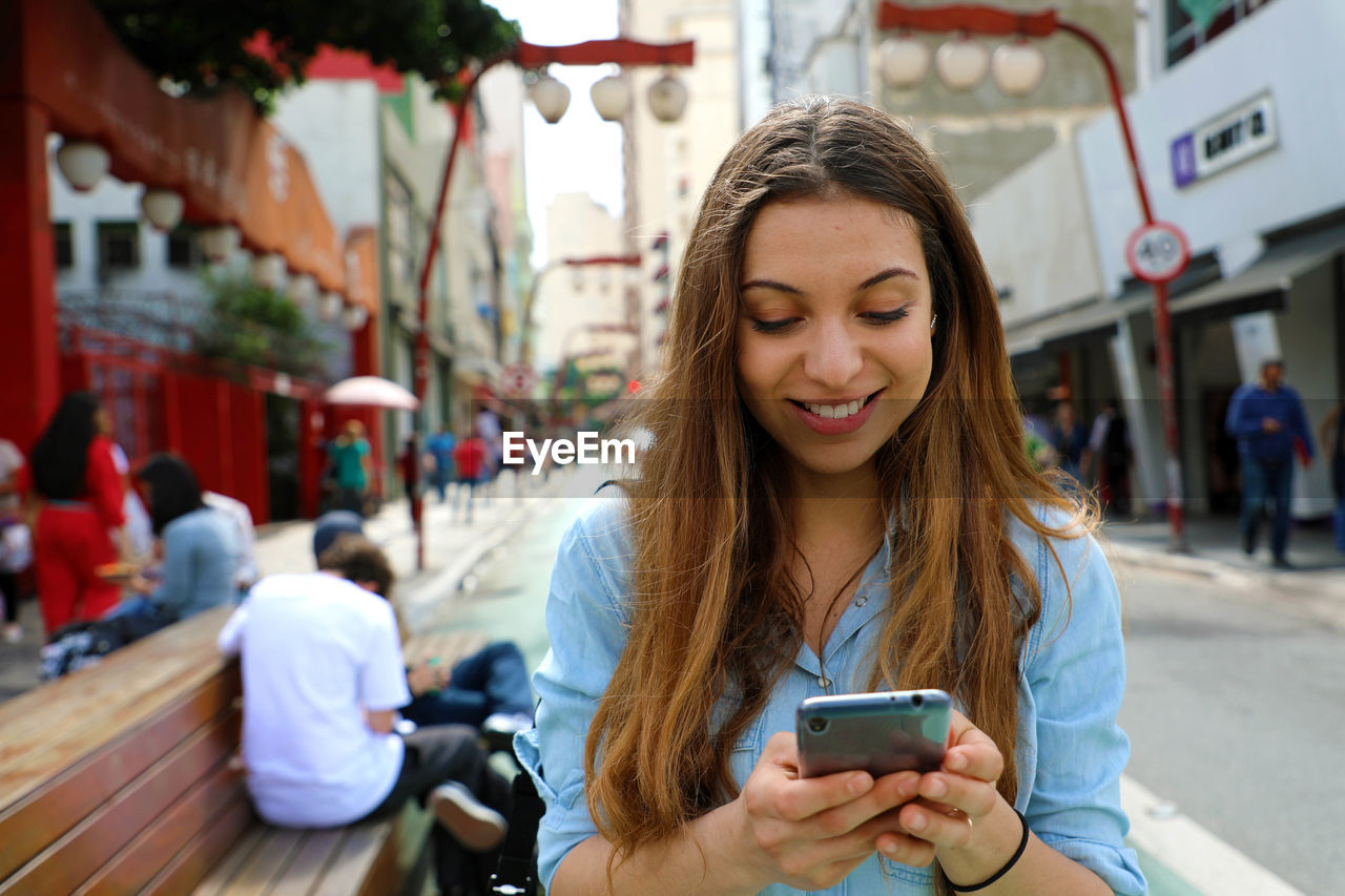 Smiling young woman using mobile phone against buildings in city