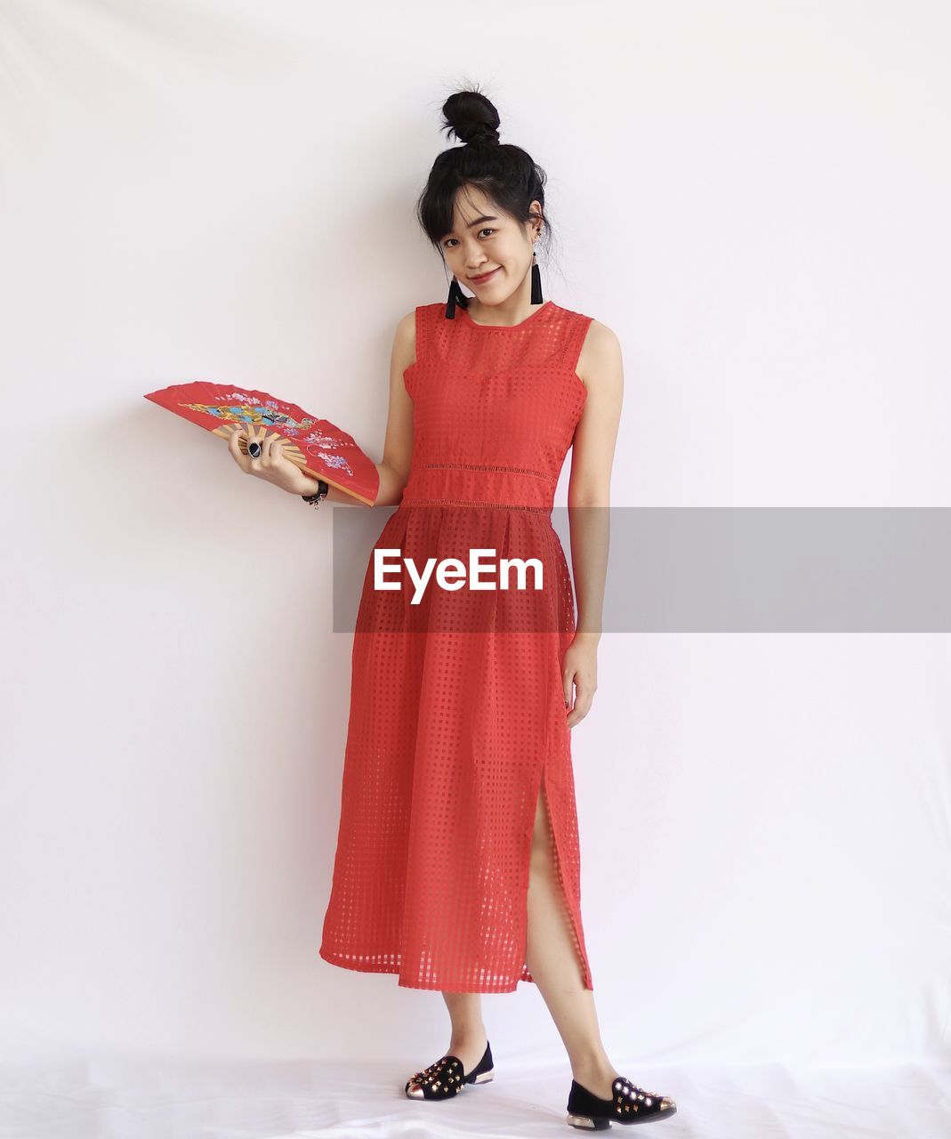 Portrait of smiling young woman in red dress holding a fan standing against white background