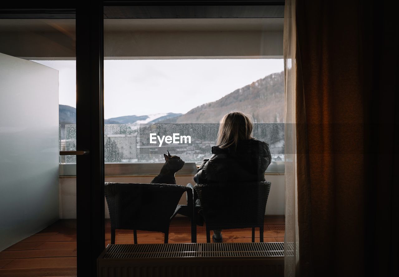 Rear view of woman sitting on chair with french bulldog in balcony by window during winter