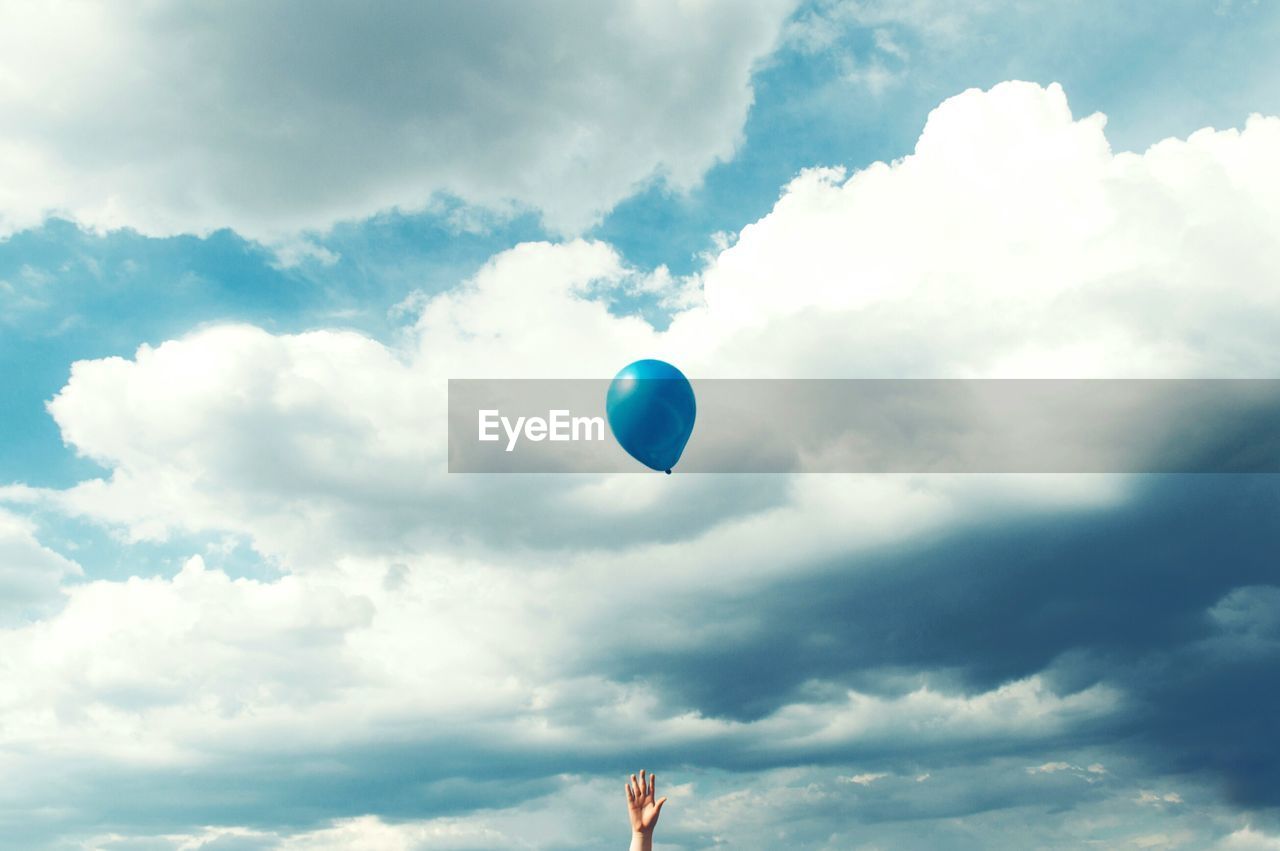 Low angle view of cropped hand with blue balloon against cloudy sky