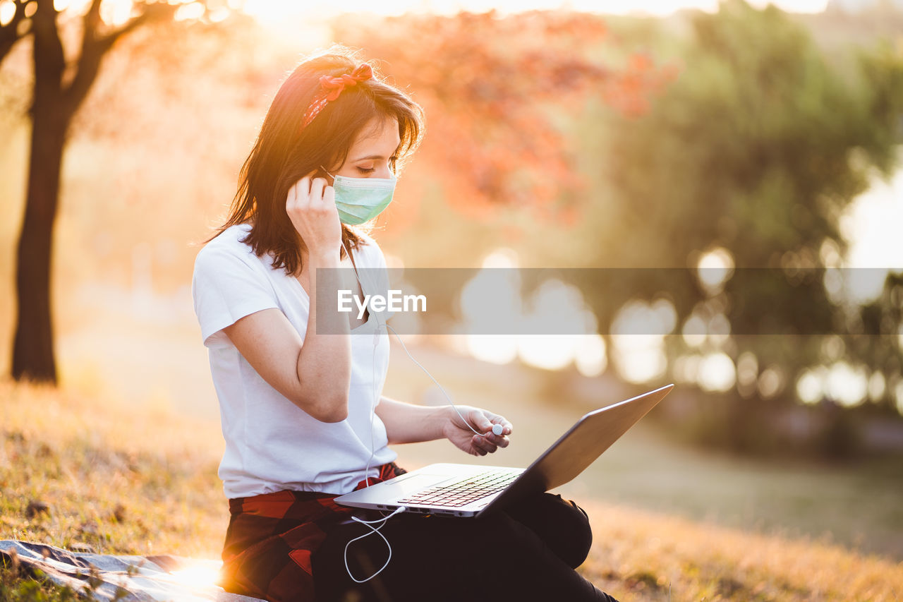 Young woman wearing mask using laptop sitting at park