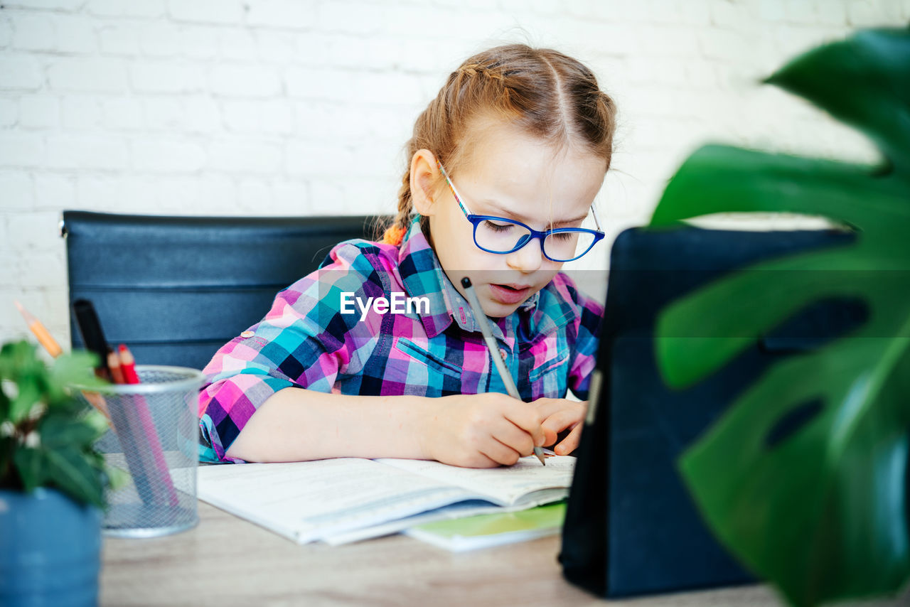 Little girl in eyeglasses making notes in her noteblook whlile studying online at home