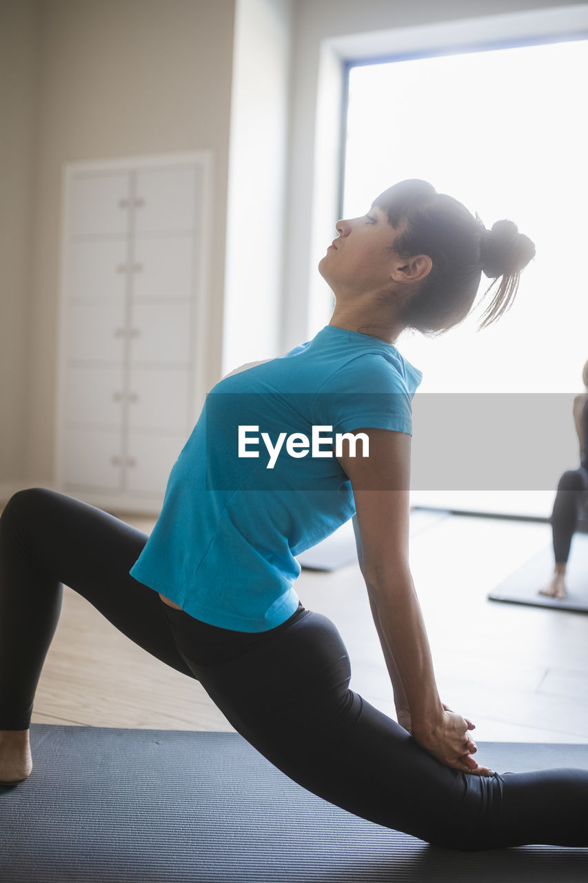 Side view of young flexible female instructor doing crescent lunge on the knee pose while practicing yoga with group of students in studio