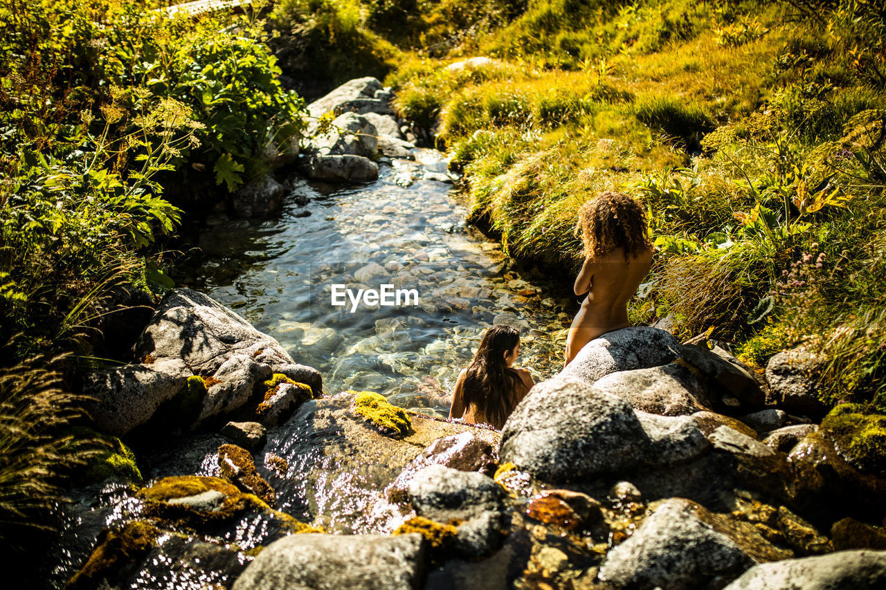From above back view of unrecognizable female travelers sitting in water of narrow stony stream flowing among green bushes in summer day in pyrenees mountains