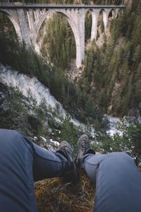 Low section of person sitting on cliff against bridge
