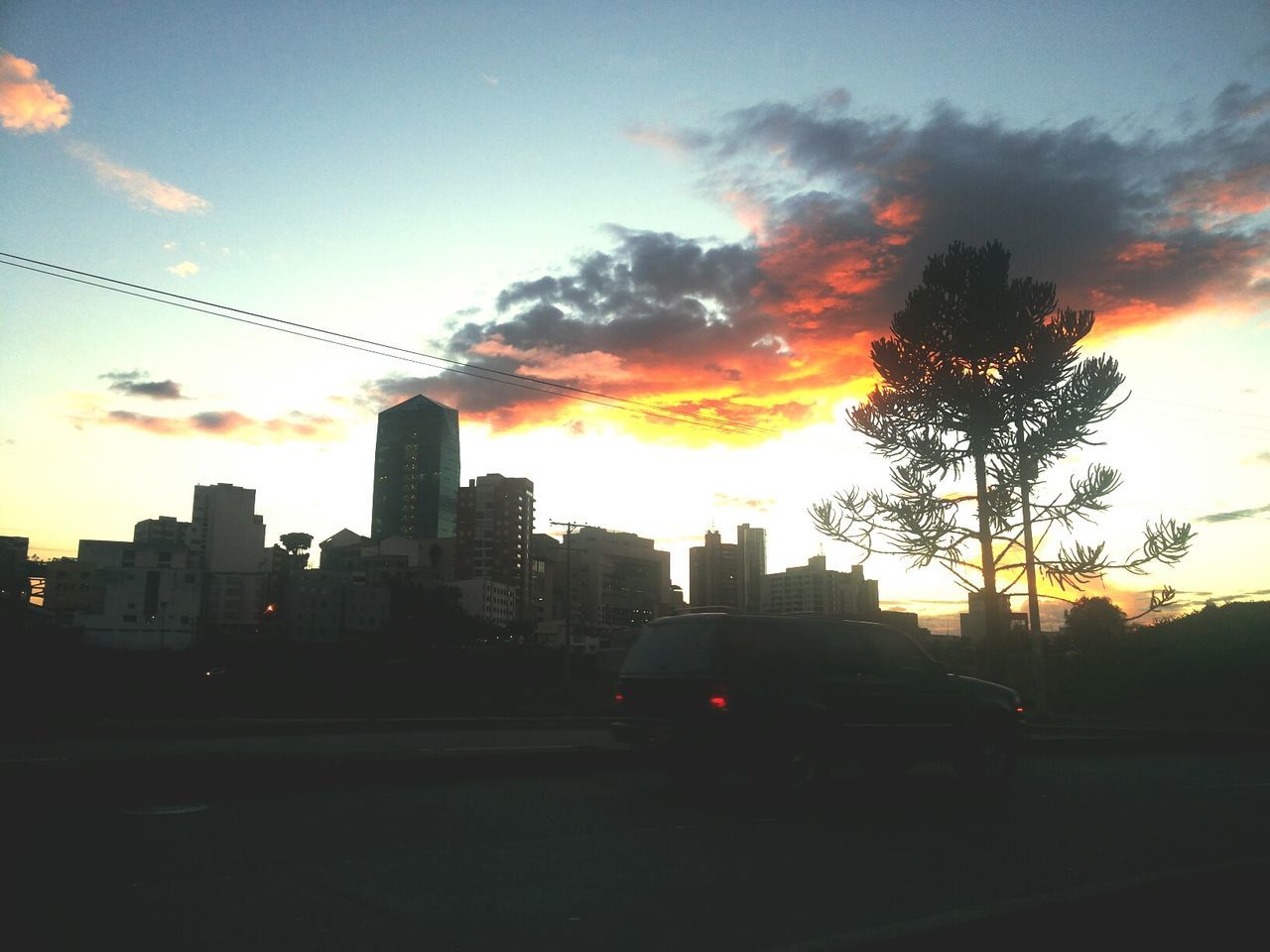 sunset, building exterior, architecture, sky, orange color, built structure, car, city, silhouette, cloud - sky, transportation, land vehicle, road, sun, street, mode of transport, cloud, cityscape, sunlight, tree