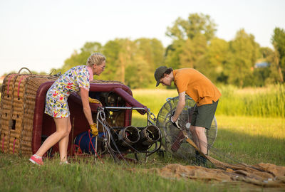 Boy assisting pilot in preparing hot air balloon on grassy field during sunset