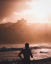 Silhouette man standing on shore against buildings during sunset