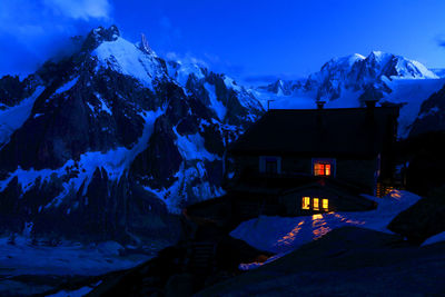 Illuminated house by snowcapped mountains against sky at dusk