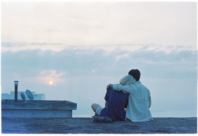 Rear view of couple sitting on terrace against cloudy sky during sunset