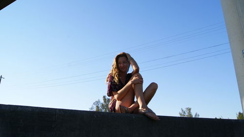 Low angle view of smiling young woman sitting on retaining wall against clear blue sky during sunny day