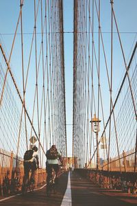 Rear view of people riding bicycles at brooklyn bridge against clear sky in city