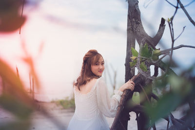 Portrait of young woman standing by tree against sky