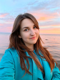 Portrait of young woman standing at beach against sky during sunset