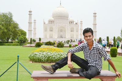 Full length portrait of young man sitting on bench against taj mahal during sunny day