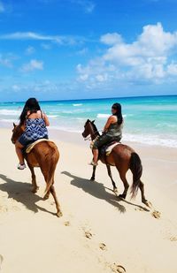 Females horseback riding at beach against sky