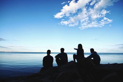 Silhouette people sitting on rock by sea against blue sky