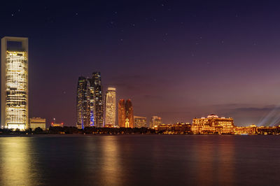 Illuminated buildings by sea against sky at night