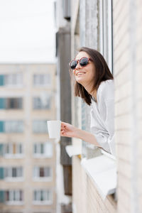 A woman in sunglasses with a cup of coffee looks out of the window and smiles. happy millennial