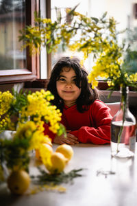 Waist up portrait of sitting at the table smiling girl with flowers in the vases in front of her