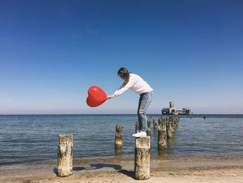 Side view of woman holding heart shape balloon on wooden post in sea against clear sky