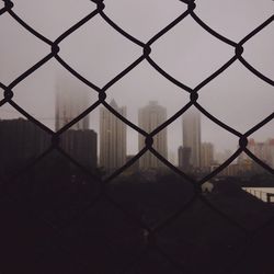 Buildings against sky during foggy weather seen through chainlink fence