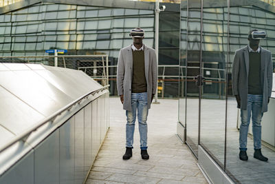 Businessman with virtual reality headset standing between glass wall and railing in city