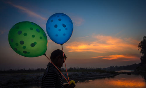 Rear view of man holding balloons against sky during sunset