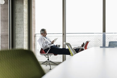 Senior manager in office sitting on chairs with feet up using digital tablet